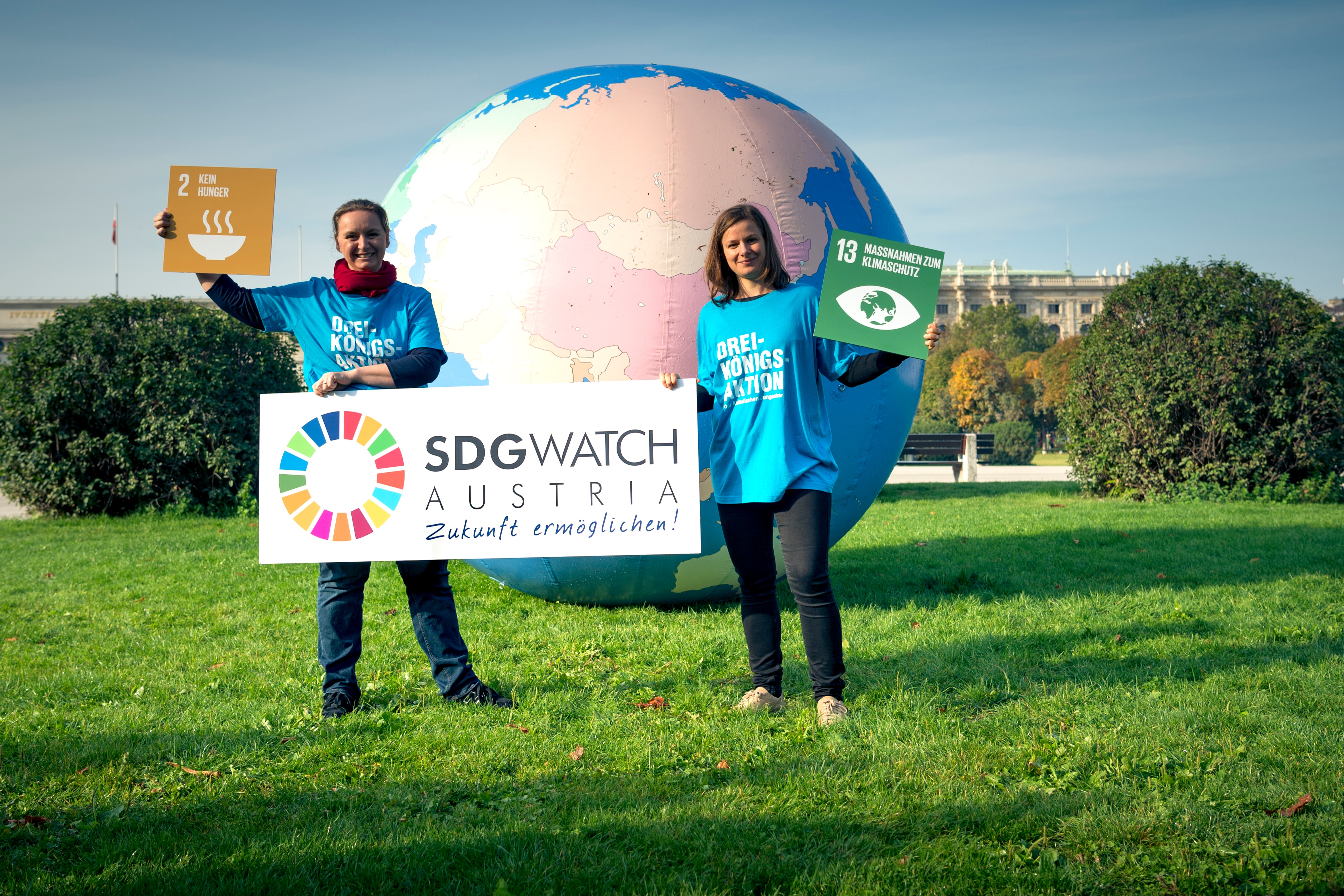 Credit: Nina Oberleitner Zwei Angestellte aus der Dreikönigsaktion halten ein Schild "SDG Watch Austria" und Schilder der SDG Ziele "2 Kein Hunger" und "13 Maßnahmen zum Klimaschutz". Hinter den beiden ist ein riesiger aufgeblasener Globus.