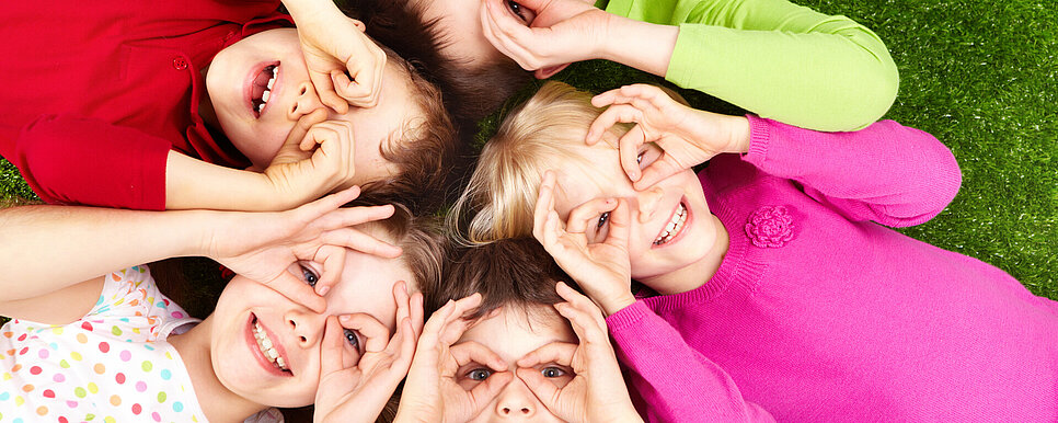 Kinder liegen auf dem Rücken im Kreis. Alle haben Daumen und Ringfinger zusammengetan und halten sie vor ihre Augen, wie eine Brille.