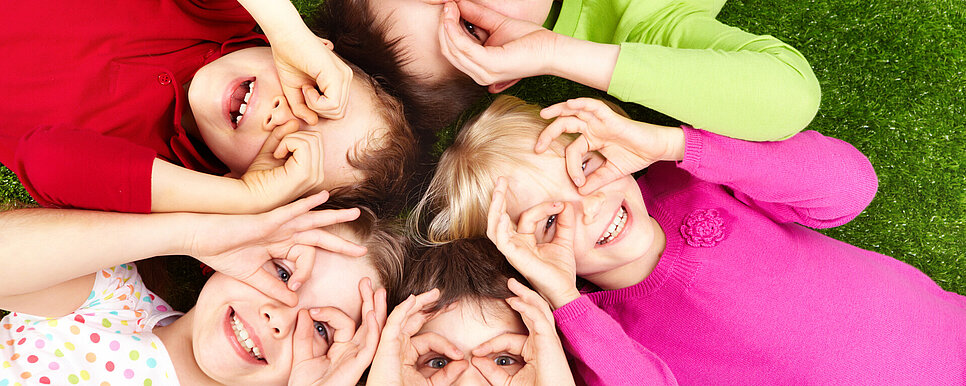 Credit: Pressmaster/shutterstock.com Kinder liegen auf dem Rücken im Kreis. Alle haben Daumen und Ringfinger zusammengetan und halten sie vor ihre Augen, wie eine Brille.