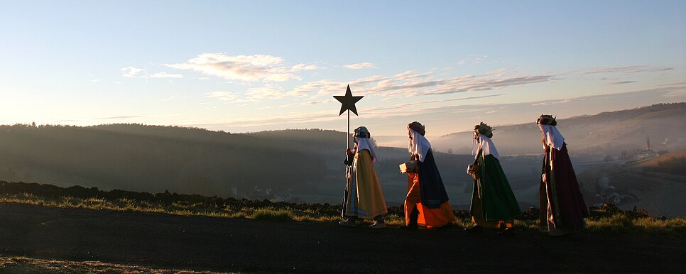 Credit: Toni Tropper Eine Sternsingergruppe geht draußen bergaufwärts auf einer Art Schotter-/ oder Wanderweg. Die vier Sternsingerkinder gehen hintereinander angeführt von dem/der Sternträger/in. Im Hintergrund hat man weiten Ausblick auf die hügelige Landschaft.