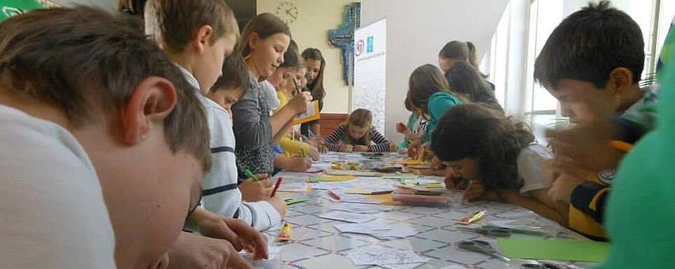 Kinder stehen rund um einen großen Tisch und schreiben oder malen.