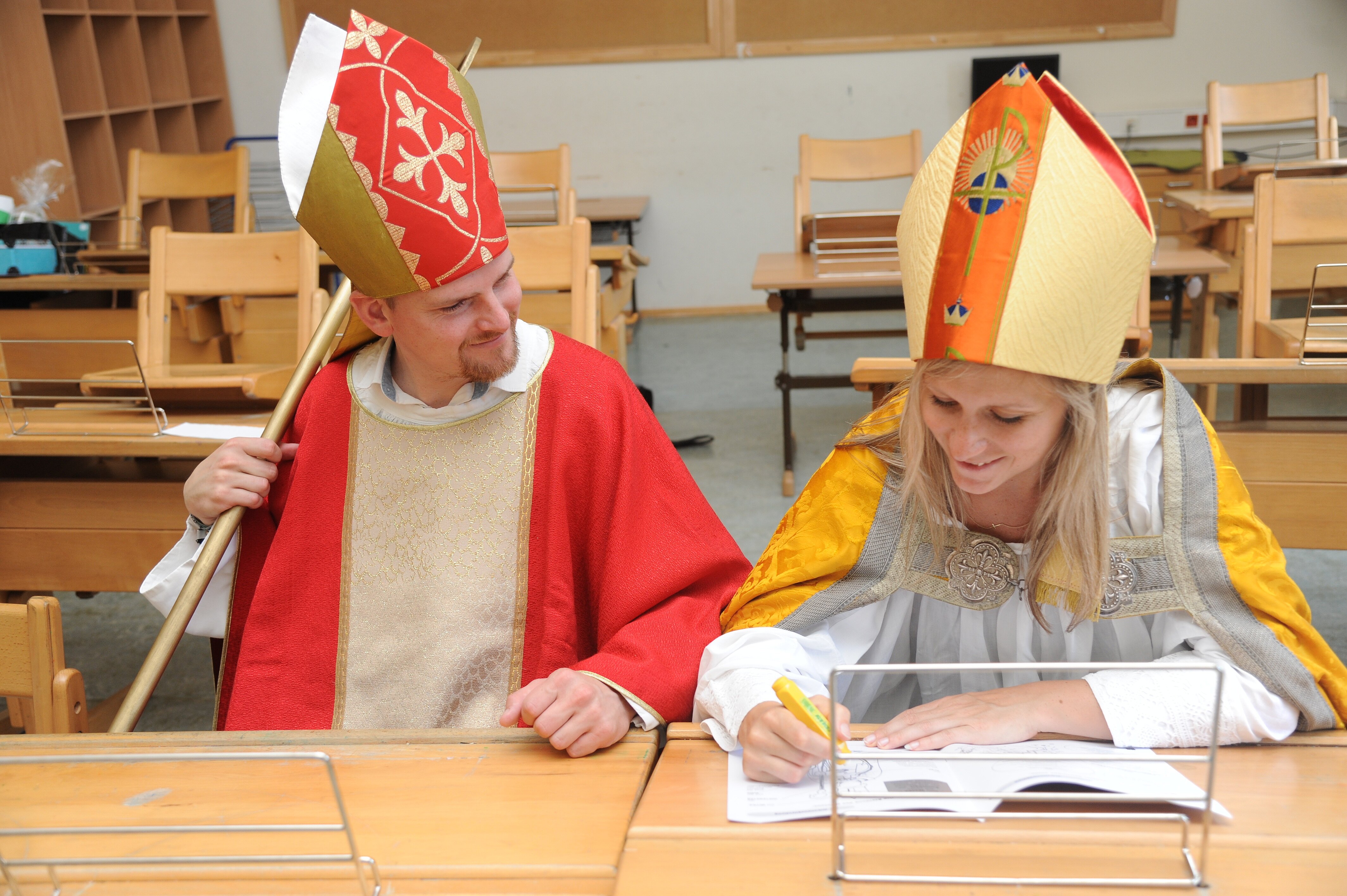 Zwei verkleidete Nikoläuse sitzen nebeneinander auf einer Schulbank in einem leeren Klassenzimmer