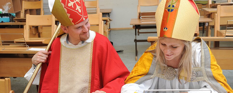 Credit: Kath. Jungschar St. Pölten Zwei verkleidete Nikoläuse sitzen nebeneinander auf einer Schulbank in einem leeren Klassenzimmer