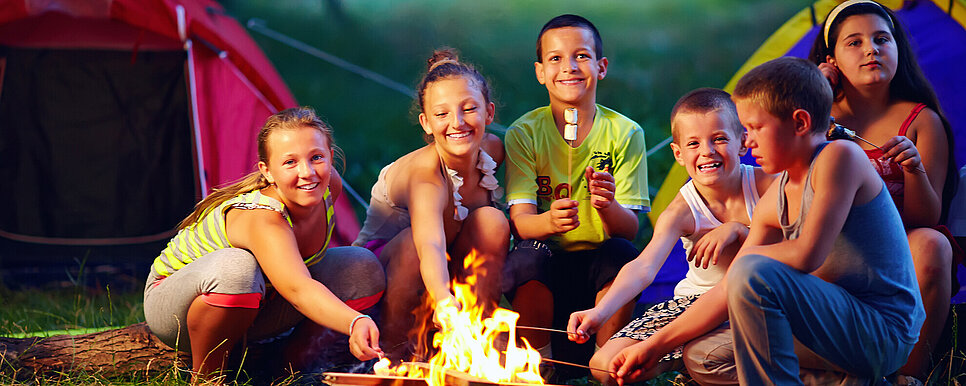 Credit: Olesia Bilkei/shutterstock.com Mehrere Kinder sitzen rund um ein Lagerfeuer und rösten Marshmallows. Dahinter sind Zelte aufgebaut.