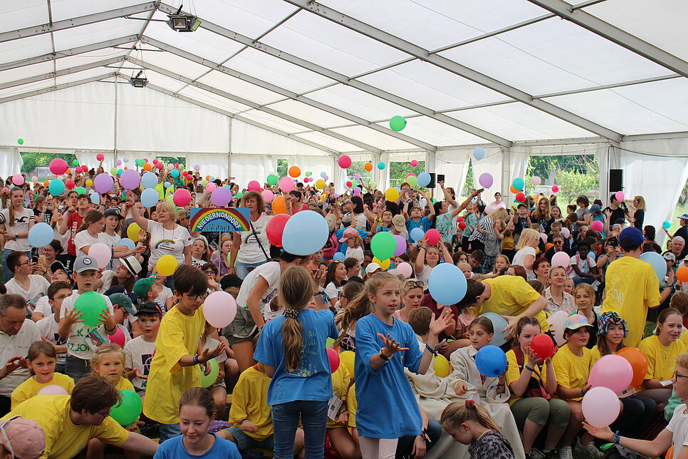 Viele Kinder sitzen und stehen in einem weißen Zelt und spielen mit Luftballons.