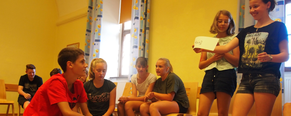 Das Bild zeigt eine Schulung für Jugendliche. Einige Jugendliche sitzen auf Sesseln im Kreis. Andere hocken bzw. stehen in der Mitte auf einem Teppich und besprechen Kärtchen auf denen einzelne Begriffe stehen.
